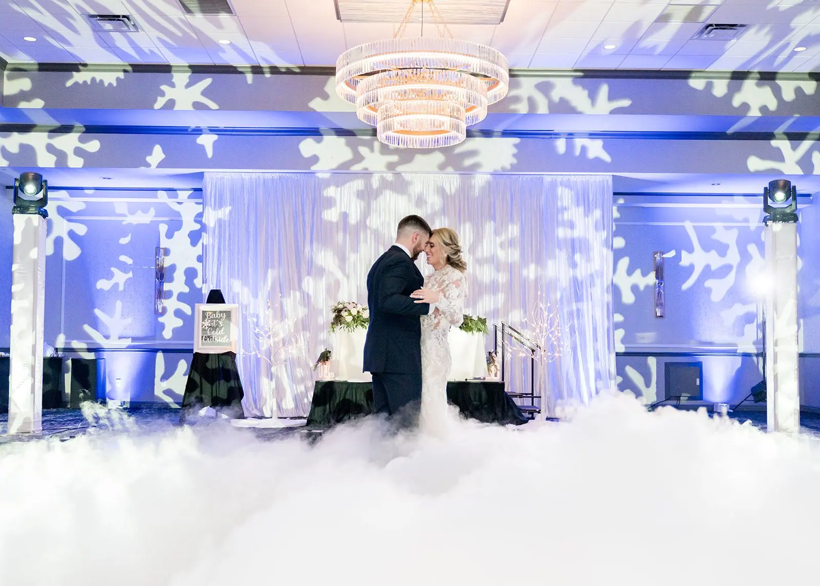 Newlyweds dancing in low-lying fog beneath snowflake lighting at a wedding reception